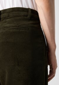 Ciemnozielone spodnie z korduroyu z pionowymi prążkami, z tylną kieszenią. Pas jest lekko podwyższony, z chropowatą powierzchnią.