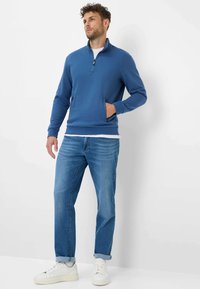 Pullover blu con zip al collo, maniche lunghe e tasche laterali, abbinato a jeans azzurri e sneakers bianche. Tessuto morbido.