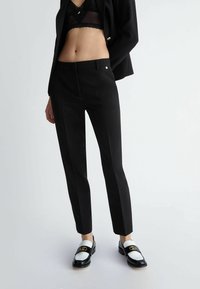 Pantaloni neri su misura con frontale piatto, passanti per cintura e vestibilità slim. Abbinati a mocassini bianchi e neri con un emblema dorato.