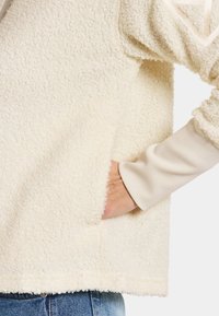 Krämfärgad fleecepolo med en texturerad yta, utrustad med en sidficka och ribbade ärmslut. Avslappnad, luftig passform.