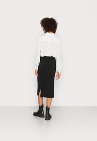 Blouse blanche à pois noirs, manches longues et col à volants, associée à une jupe noire longueur genou avec une fente latérale. Bottines noires à la cheville.