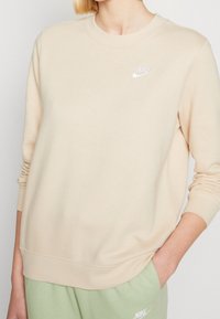 Beige Nike sweatshirt med rund hals och ribbade manschetter, med en vit logotyp på bröstet. Mjuk, slät tygtextur.