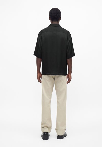 Homme debout, dos à nous, portant une chemise noire ample à manches courtes, un pantalon beige et des chaussures noires sur fond blanc.