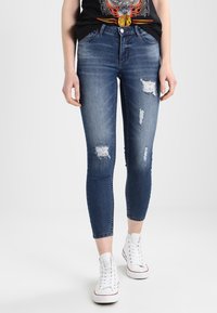 Persona vistiendo jeans ajustados rasgados de color azul oscuro, camiseta gráfica negra y zapatillas altas blancas, de pie sobre un fondo blanco.