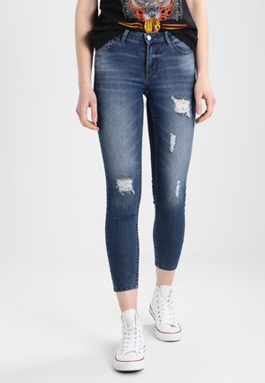 Persona vistiendo jeans ajustados rasgados de color azul oscuro, camiseta gráfica negra y zapatillas altas blancas, de pie sobre un fondo blanco.