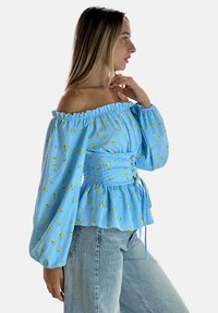 Niebieska bluzka typu off-shoulder z żółtym kwiatowym wzorem, marszczoną talią, długimi bufkowymi rękawami i detalem w postaci wiązania. W zestawie z jasnymi jeansami.