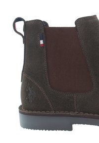 Bota de tobillo de ante marrón con paneles laterales elásticos y una parte trasera texturizada en marrón. Presenta un detalle bordado y un pequeño logo.