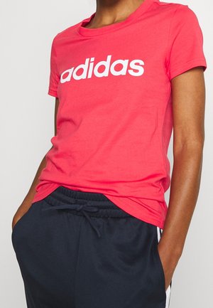 Osoba nosząca różowy t-shirt Adidas z białym logo i ciemnoniebieskie spodnie ze sznurkiem, ręce w kieszeniach na jednolitym tle.