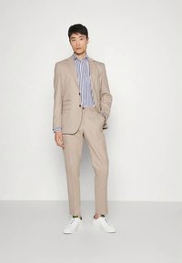 Beżowy garnitur z marynarką na dwa guziki i spodniami o prostych nogawkach, w połączeniu z niebiesko-białą koszulą w paski oraz białymi trampkami.