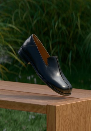Black leather loafer shoe balancing on the edge of a wooden table with green foliage in the background.