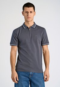 Graues Poloshirt mit Reißverschluss am Kragen, kontrastierenden weißen Streifen an den Ärmeln, kurzen Ärmeln und leicht texturierter Stoff. Wird mit blauer Jeans getragen.