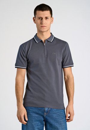 Polo grigia con collo con zip, strisce bianche a contrasto sulle maniche, maniche corte e tessuto leggermente strutturato. Indossata con jeans blu.