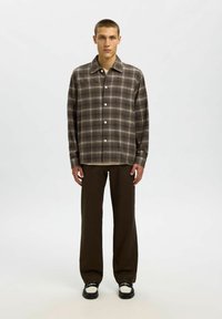 Jauns vīrietis stāv, ģērbies brūnā raibā krekla ar pogām, tumši brūnās biksēs un melnbaltos mokasīnos uz viendabīga gaiša fona.