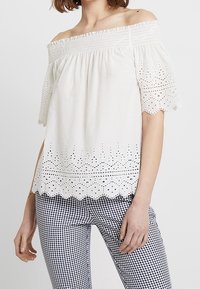 Top blanco con escote off-the-shoulder y mangas cortas abullonadas, con detalles de encaje de ojo de perdiz y un dobladillo con forma de olas. Combinado con pantalones de patrón de cuadro Vichy.