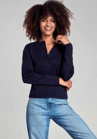 Cardigan bleu marine avec un col rond, des finitions côtelées et des boutons, associé à un jean bleu clair. Texture douce, coupe ajustée, style polyvalent.