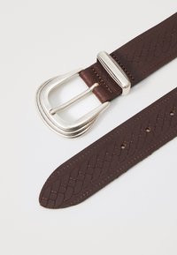 Curea din piele maro cu o textură împletită, cataramă din metal argintiu cu o formă curbată și mai multe găuri uniform distanțate pe curea.