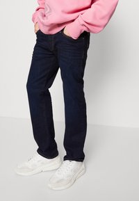Jeans de mezclilla azul oscuro con corte recto, combinados con zapatillas blancas de malla. Un sudadera rosa está puesta encima, con las manos en los bolsillos.