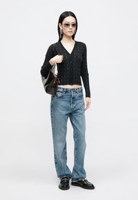 Zwarte gebreide cardigan met een v-hals en knopen, gecombineerd met blauwe relaxed-fit jeans. Accessoires zijn onder andere een zonnebril en een bruine schoudertas.