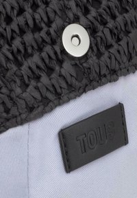 Textil negro tejido con acabado texturizado, que presenta un cierre de broche plateado y una etiqueta de cuero negro con el grabado "TOUS". Base de tela blanca.