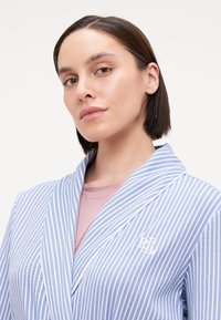 Ljusblå randig morgonrock med vita detaljer och logotyp. Har en sjalkrage och skräddarsydd passform. Slät tygtextur, lämplig för avkopplande kläder.