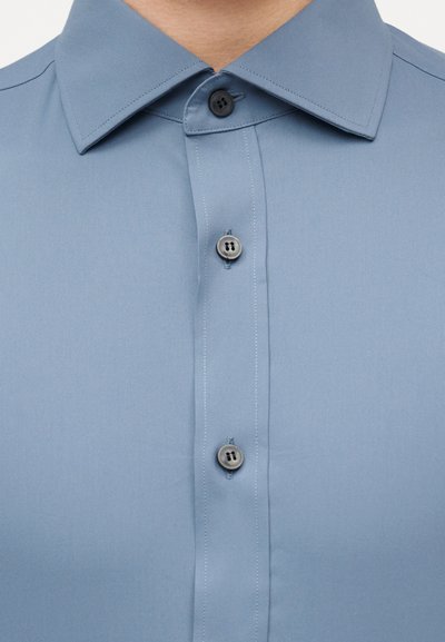 Κοντινό πλάνο ενός ατόμου που φοράει ένα γαλάζιο πουκάμισο με κουμπιά και μυτερό γιακά.