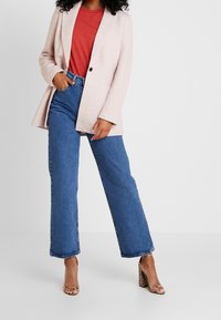 Jasnoróżowa marynarka o dopasowanym kroju na czerwonej koszulce, zestawiona z szerokimi niebieskimi dżinsami i wzorzystymi sandałami na słupku.