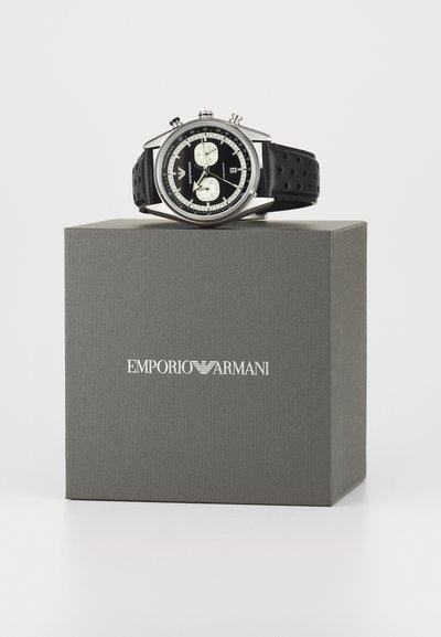 Sort chronografur med sølv detaljer, rustfrit stål urkasse og perforeret gummirem, placeret på en grå æske mærket "Emporio Armani."