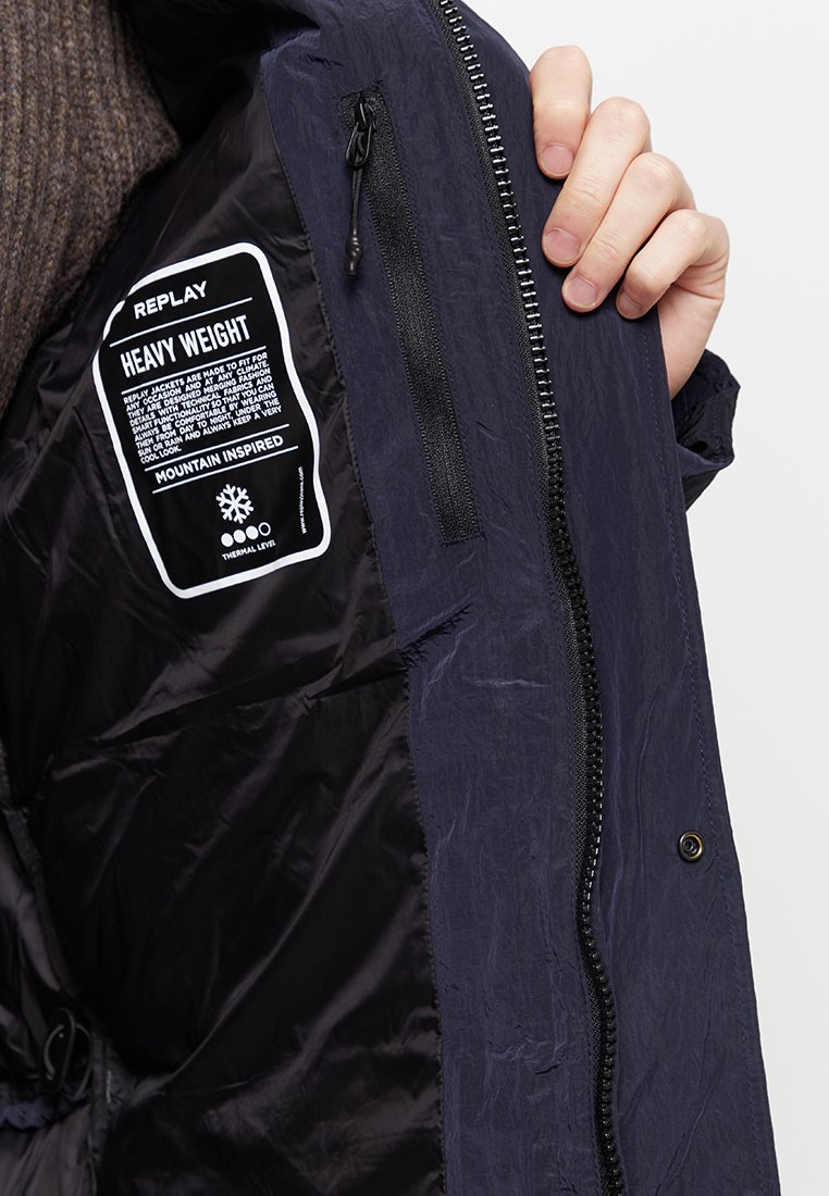 Binnenkant van een marineblauwe gewatteerde jas met een zwaar label; heeft een ritssluiting en een gestructureerde stof met een gladde, glanzende voering.
