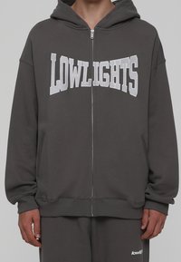 Sudadera gris con cremallera y la palabra "LOWLIGHTS" en letras grandes blancas, hecha de material suave, con capucha y bolsillos laterales.