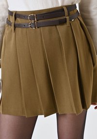 Gonna plissè khaki con due cinture marroni scure dotate di fibbie dorate. Il tessuto ha una texture liscia e una silhouette sartoriale.