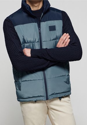 Hombre con un chaleco acolchado azul y azul marino sobre un jersey negro de punto con los brazos cruzados, pantalones beige visibles desde la cintura hacia abajo.