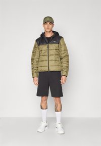 Giacca puffer verde oliva con parte superiore nera, zip frontale e cappuccio. Abbinata a pantaloni corti neri, calze bianche e sneakers bianche.