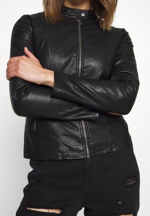 Personne portant une veste en cuir noire avec une fermeture éclair à l’avant et un jean noir déchiré, bras croisés, cadrée au niveau du cou et des cuisses.