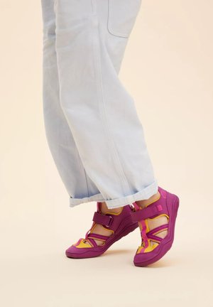 Purple and yellow sandals with Velcro straps, a textured sole, and open design, paired with light blue cuffed jeans.