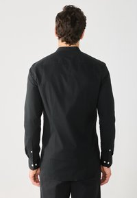 Maglietta nera a maniche lunghe in cotone con orlo curvo, colletto standard e bottoni a pressione argentati sui polsini. Texture liscia e design aderente.