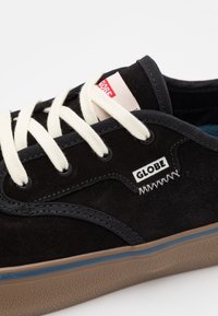 Baskets en daim noir avec lacets blancs, semelle beige, et logo "Globe" sur le côté, vue rapprochée de la tige et des œillets.