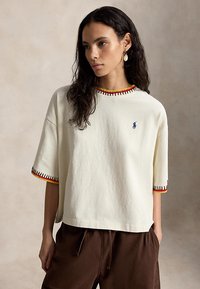 Kvinne med mørkt bølget hår iført kremfarget, oversized kortermet skjorte med flerfarget kant og brune bukser, stående mot ensfarget bakgrunn.