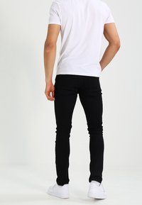 Svarta smala jeans med en slät textur, bärs med en vit t-shirt och vita sneakers, som visar en åtsittande silhuett bakifrån.