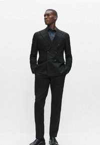 Sort dobbeltradet jakkesæt med skræddersyet pasform, med hakkede revers og to frontlommer. Kombineret med en mørk skjorte og skinnende sorte sko.