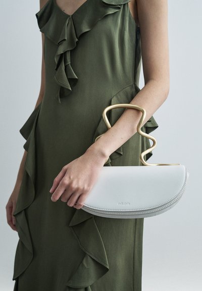 Kvind i olivengrøn, voluminøs kjole, som holder en hvid halvmåne-formet taske med et abstrakt guldhåndtag foran en ensfarvet baggrund.