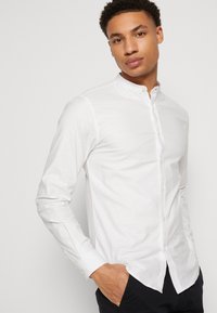Camisa blanca de manga larga con cuello mao, hecha de una tela ligera, que presenta una textura suave y un diseño frontal liso.