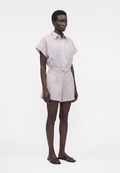 Γυναίκα που φοράει ριγέ ολόσωμη φόρμα με κοντά μανίκια, κουμπιά, κορδόνι στη μέση και μαύρες σαγιονάρες, στέκεται μπροστά σε λευκό φόντο.