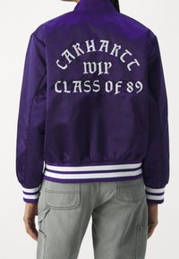 Paarse satijnen bomberjack met witte tekst "CARHARTT WIP CLASS OF 89" op de achterkant, voorzien van gestreepte witte en paarse ribbels aan de mouwen en onderkant.