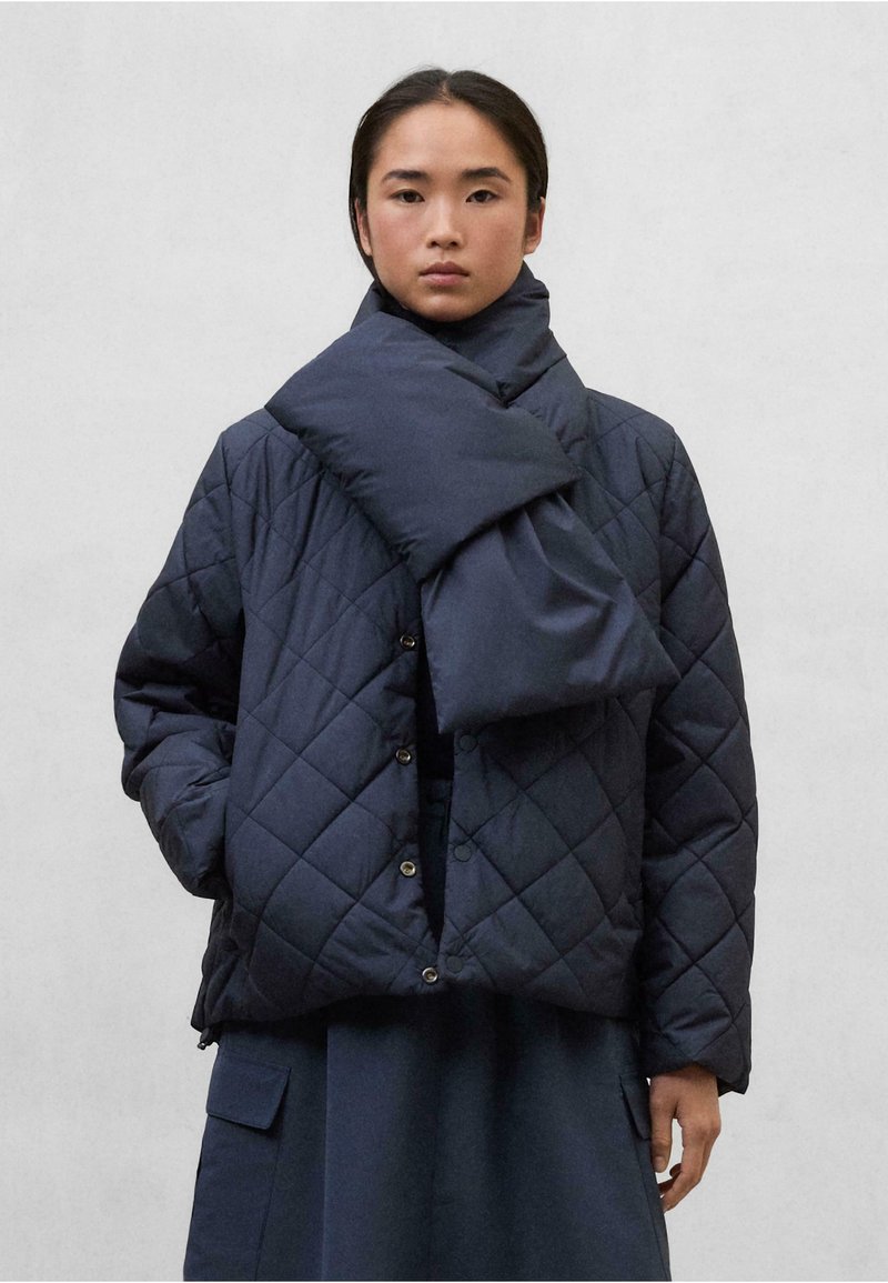 Chaqueta acolchada en color marino con cuello alto y cierre de botones, que presenta una bufanda a juego colocada sobre los hombros. Tejido texturizado, ajuste holgado.