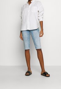 Camisa blanca de algodón con botones y un bolsillo en la parte frontal, combinada con pantalones cortos de mezclilla azul claro hasta la rodilla y sandalias negras tipo deslizante.