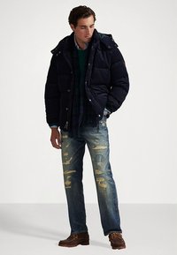 Marineblauw donzen jack met capuchon, blauw geruite sjaal, groene trui, versleten blauwe spijkerbroek en bruine veterlaarzen. Opvallende slijtagepatronen op de spijkerbroek.