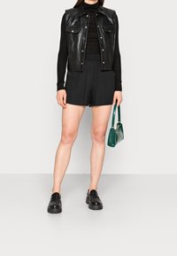 Schwarze Lederweste über einem schwarzen Rollkragenpullover, kombiniert mit schwarzen Shorts. Mühelose schwarze Schuhe und eine grüne Tasche mit Krokoprägung runden den Look ab.