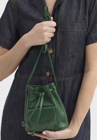 Sac seau en cuir vert à cordon de serrage avec une forme arrondie, une texture lisse, une poche avant et une sangle réglable tenue dans une main.