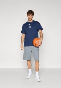 Námornícky modré bavlnené tričko s bielym logom, šedé šortky a biele tenisky. Osoba drží basketbal. Jednoduchý dizajn, voľný strih.