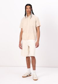 Camisa de manga corta beige con dos bolsillos frontales, combinada con pantalones cortos crema. El modelo lleva zapatillas blancas y calcetines blancos. Tejido de textura suave.